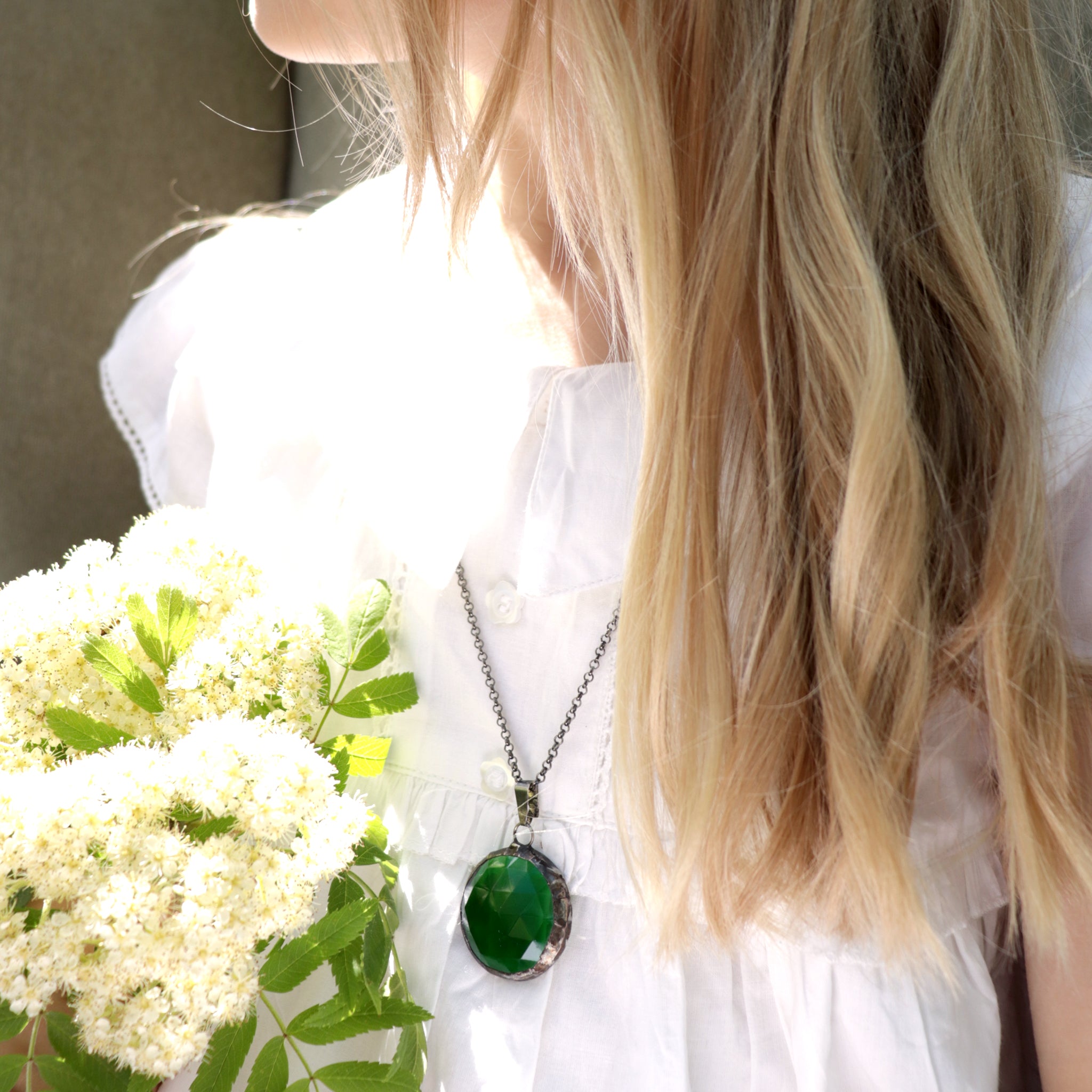 Blonde girl in white dress wearing emerald necklace holding bunch of flowers