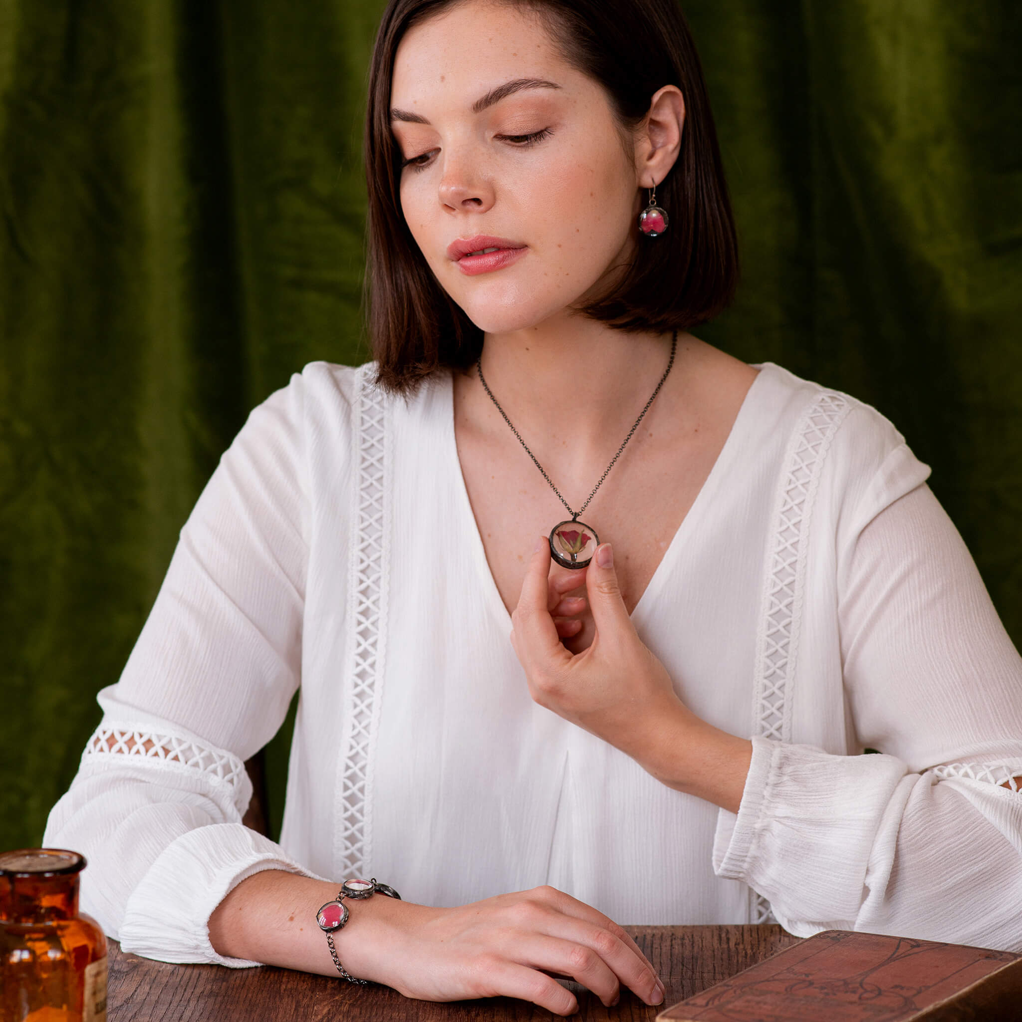 Brunette in a white blouse sitting by the table holding hand on her rose necklace