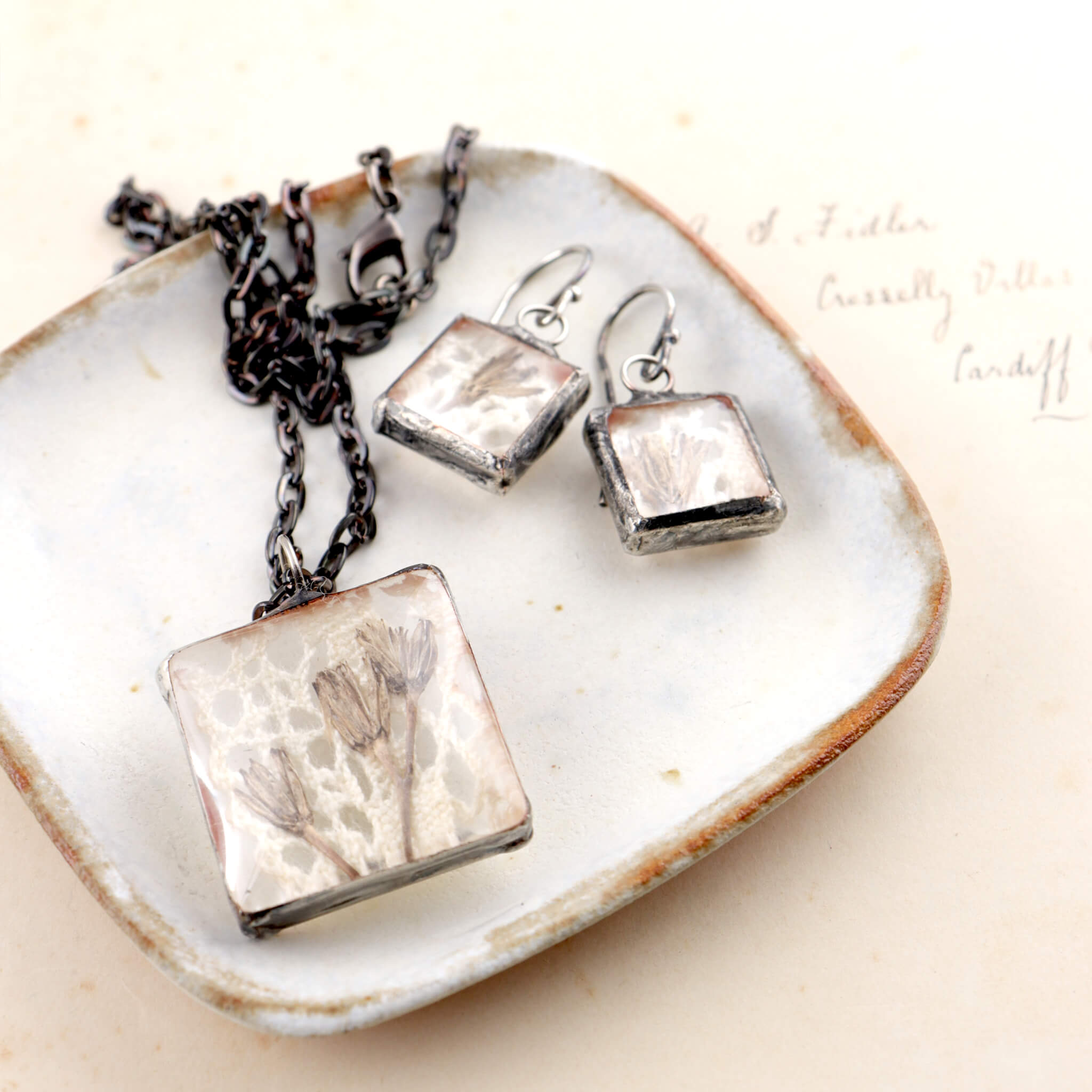 Square soldered jewellery set with vintage lase and pressed flowers lying on a shite dish