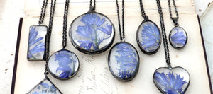 pressed bluebell flowers framed into glass soldered necklaces in variety of shapes lying side by side in line