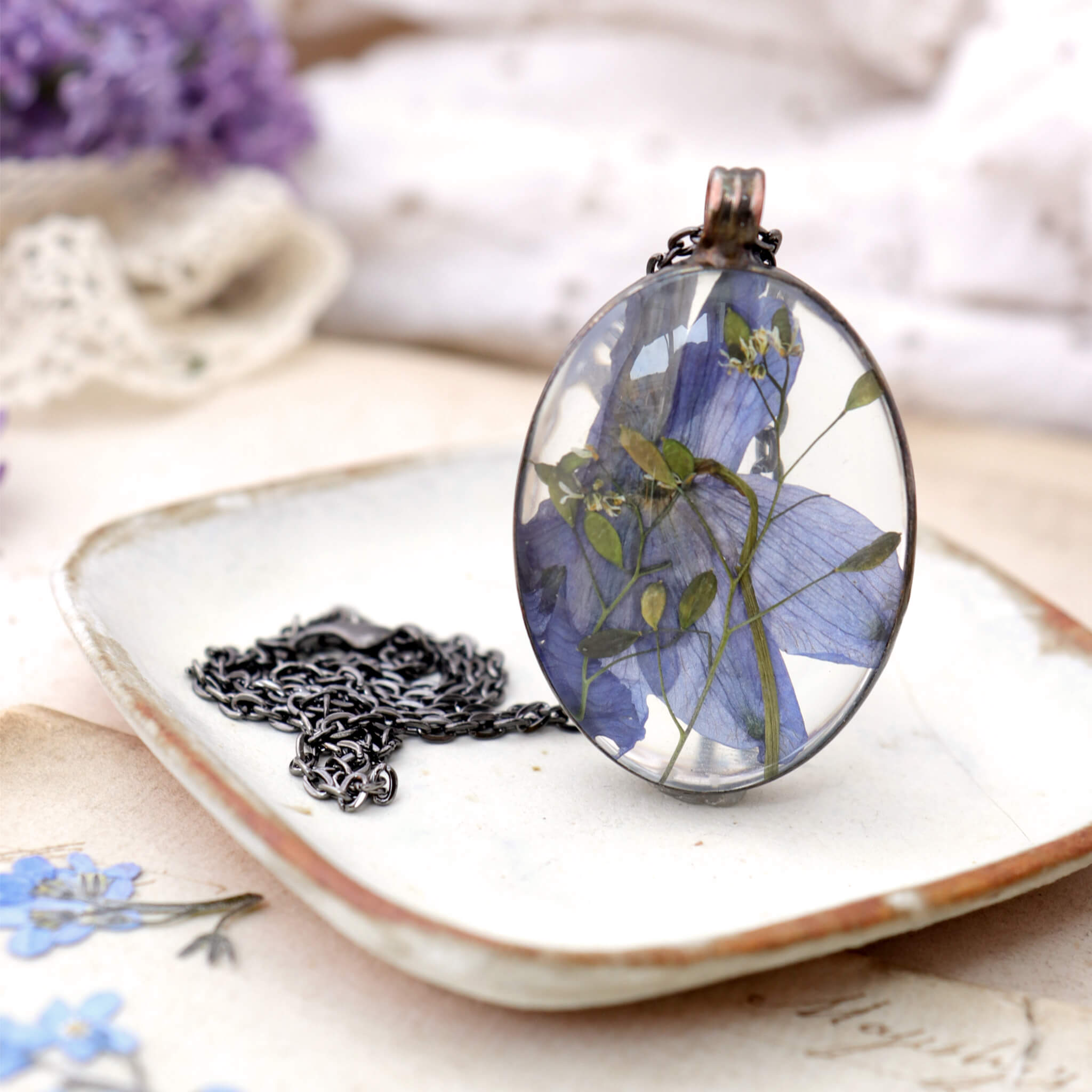 Blue pressed flower in a glass soldered necklace standing up on a white dish