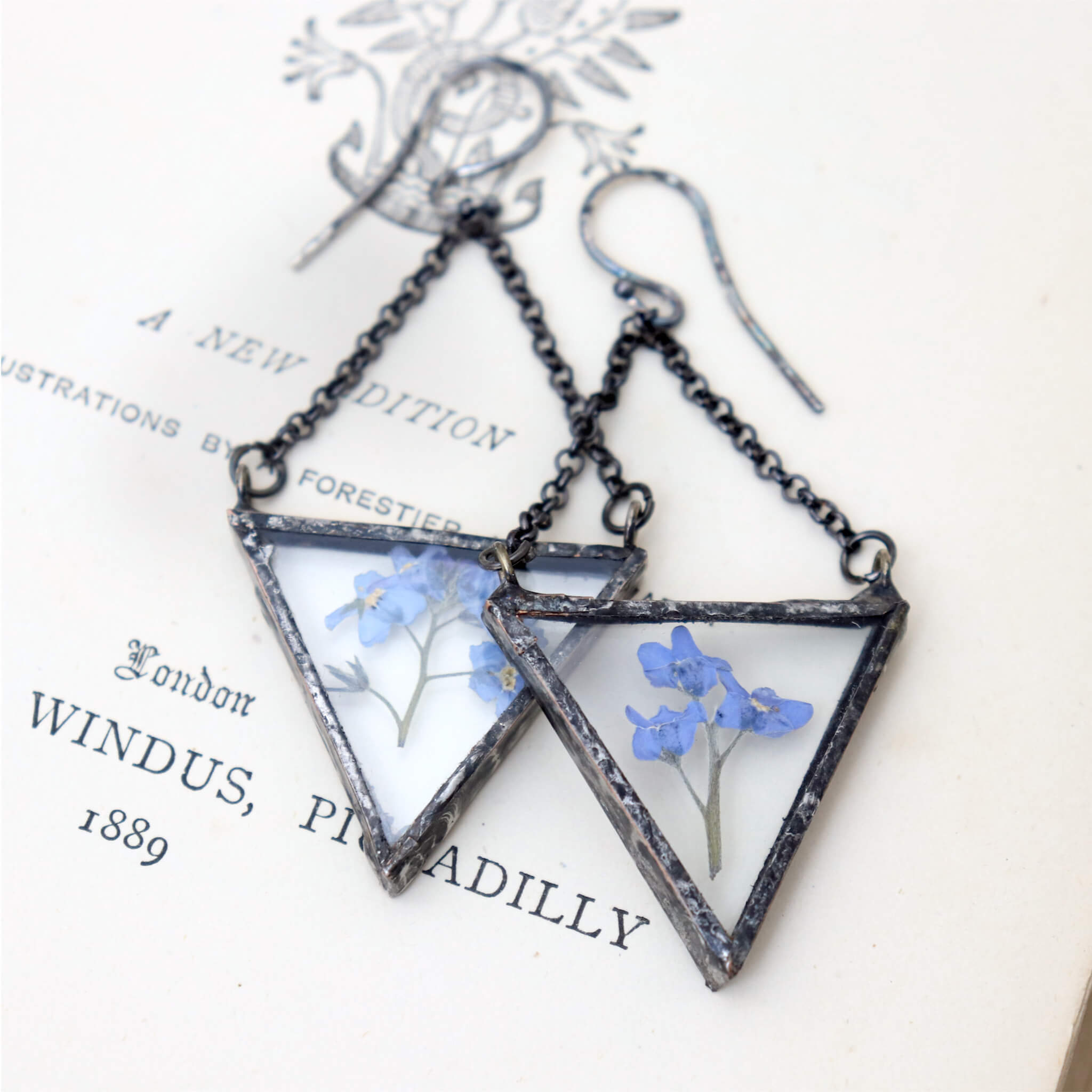 Triangular pressed blue forget-me-not flowers earrings lying on an old book