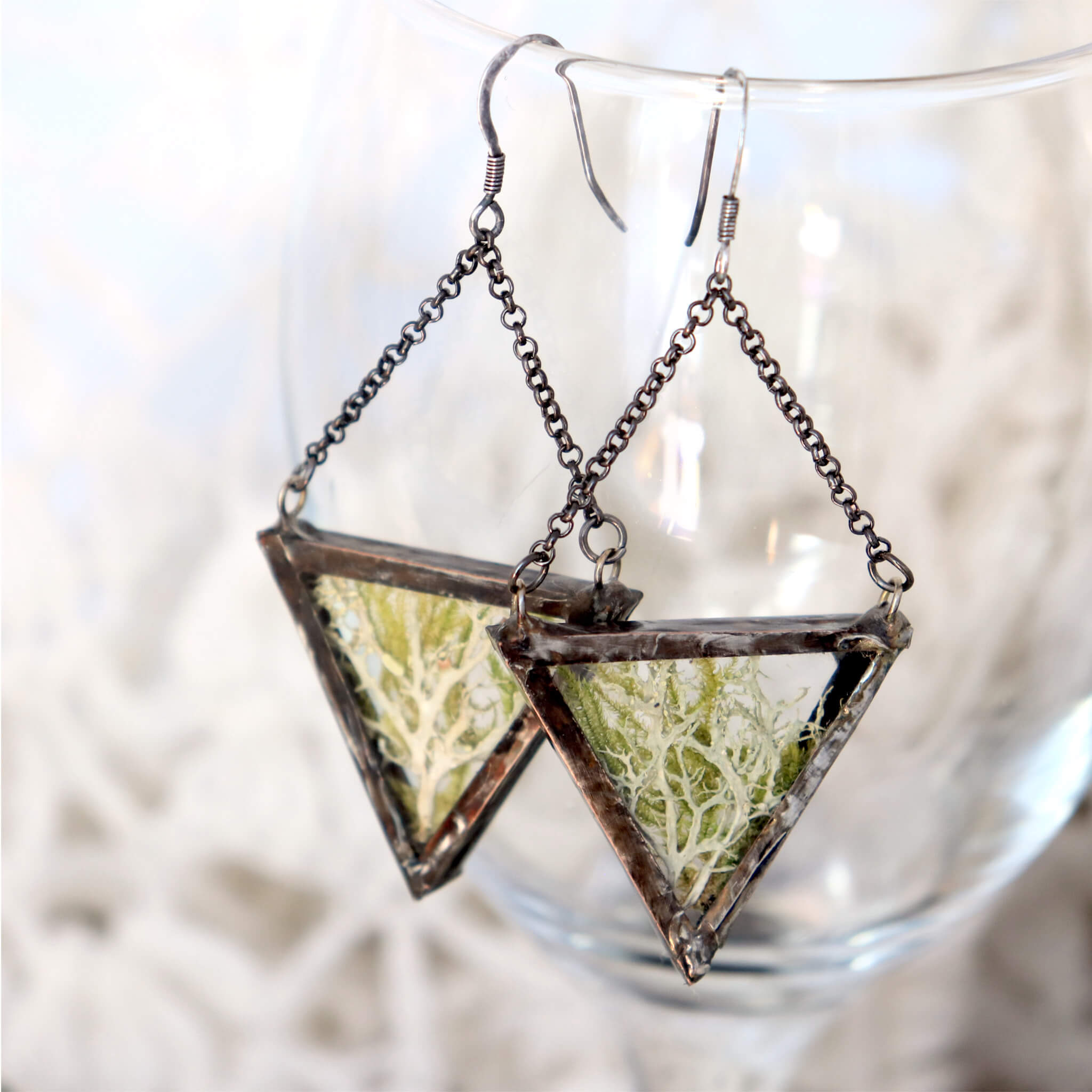 Triangular pressed moss and lichen earrings hanging from an edge of a glass
