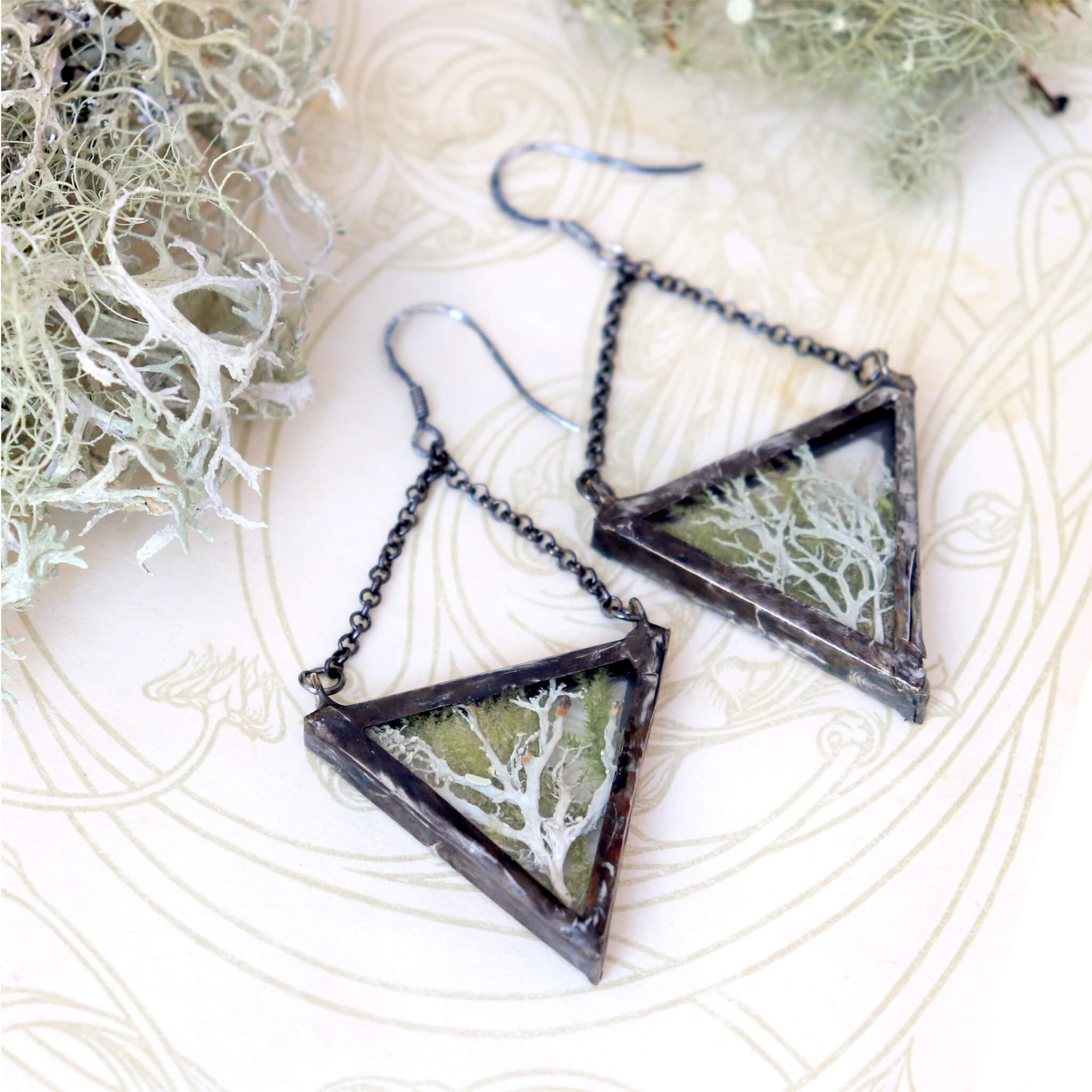 Triangular pressed moss and lichen earrings lying on an old book
