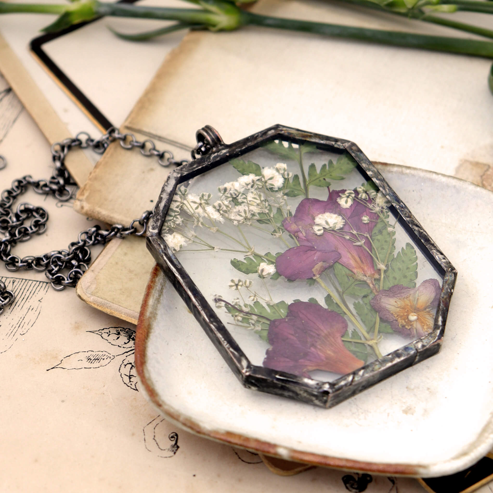 pink flowers and green ferns in glass and solder necklace lying on a white dish