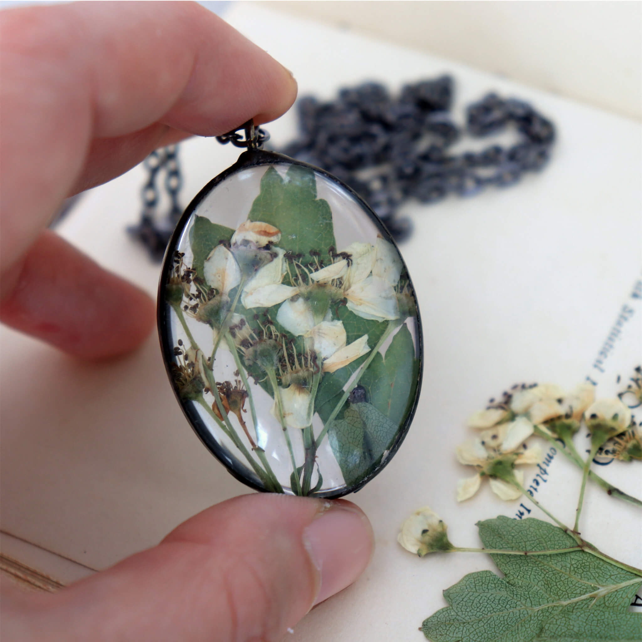 Hand holding a oval shaped glass necklace with hawthorn flowers inside