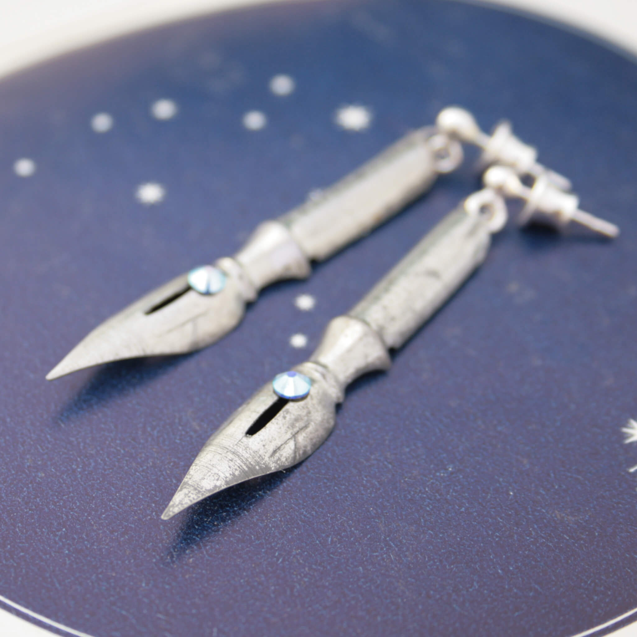 pen nib earrings with aquamarine birthstone crystals on them lying on a constellation print