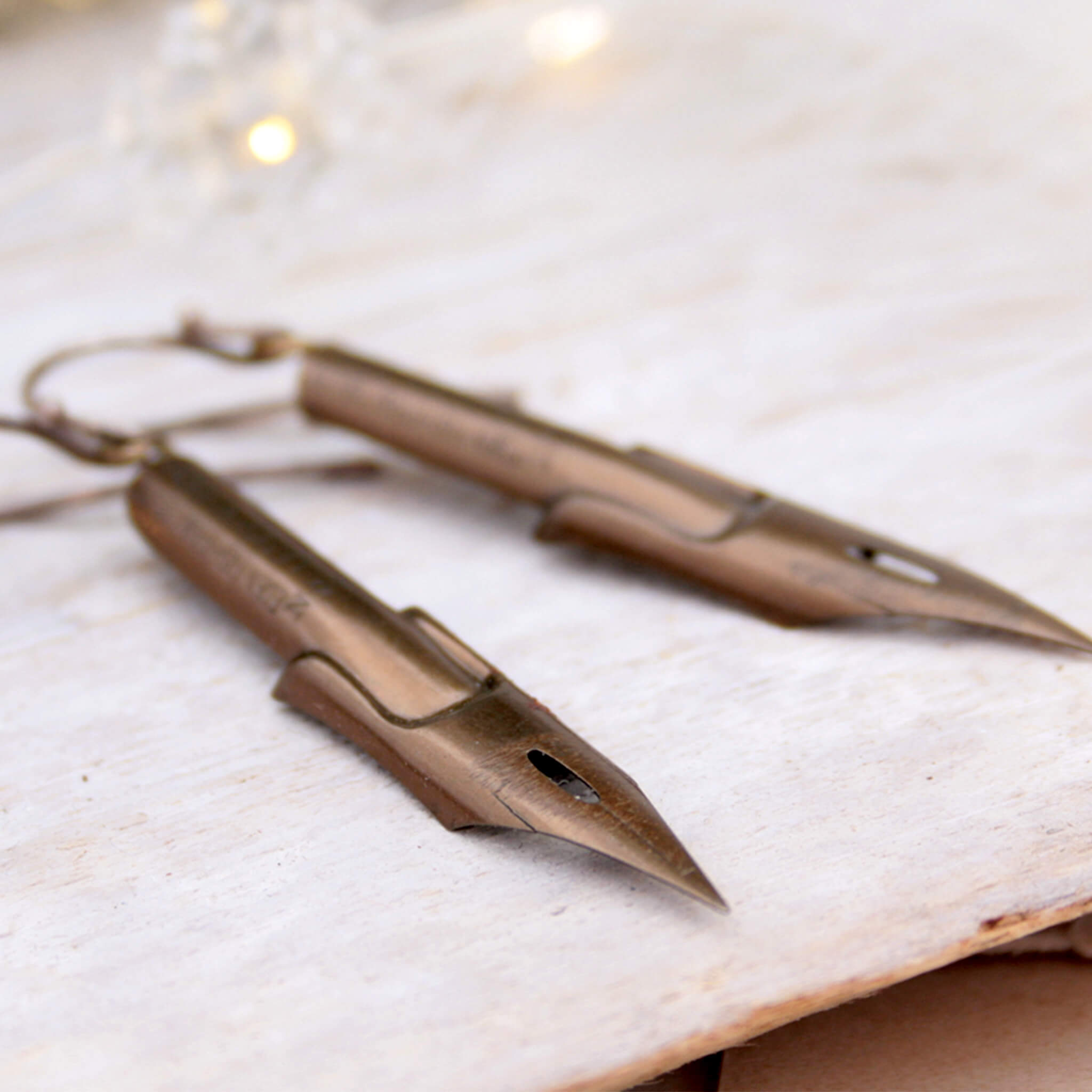 dark academia earrings made of pen nibs in bronze colour lying on a wooden plank painted white