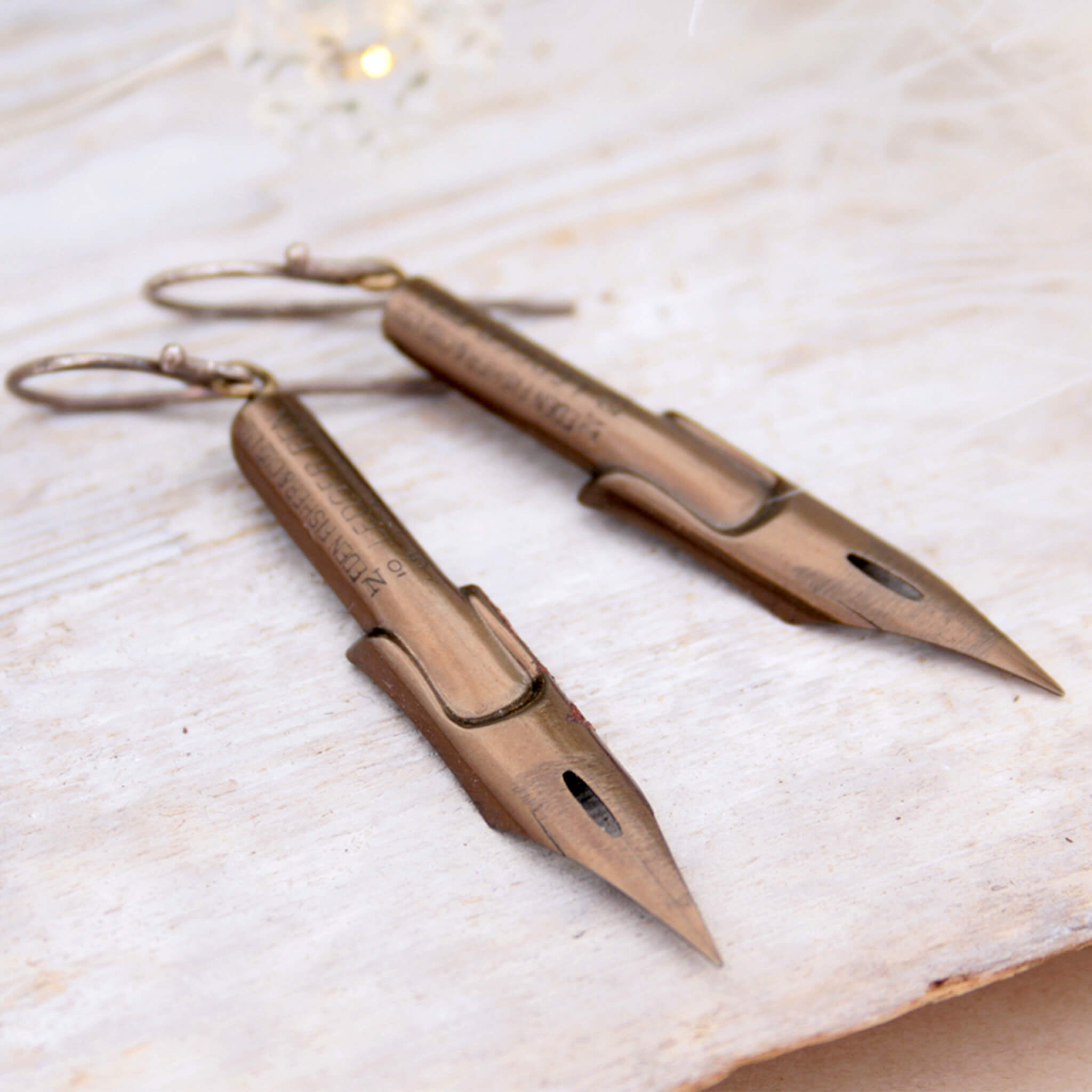 dark academia earrings made of pen nibs in bronze colour lying on a wooden plank painted white