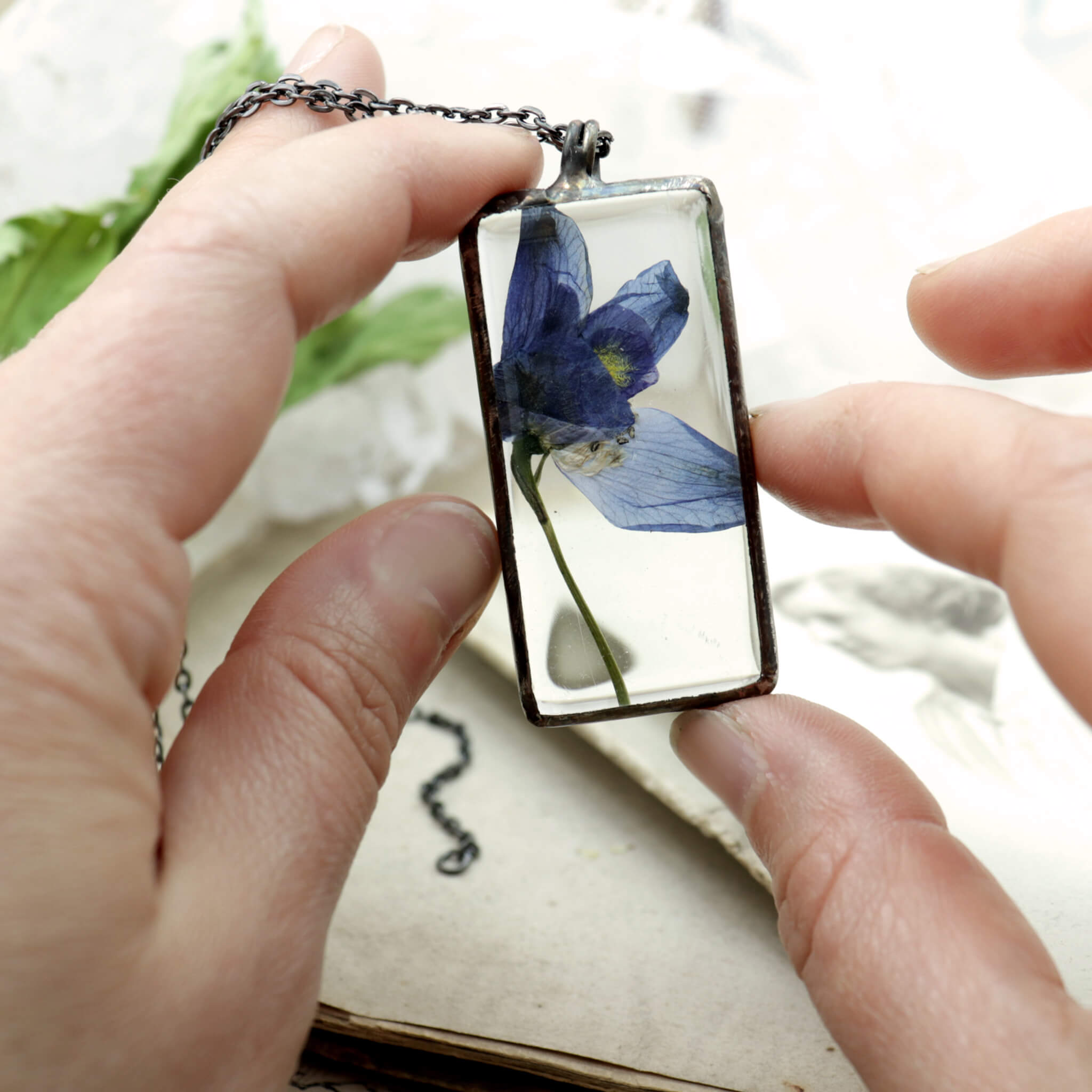 Hands holding a blue pressed flower in a rectangular glass soldered necklace