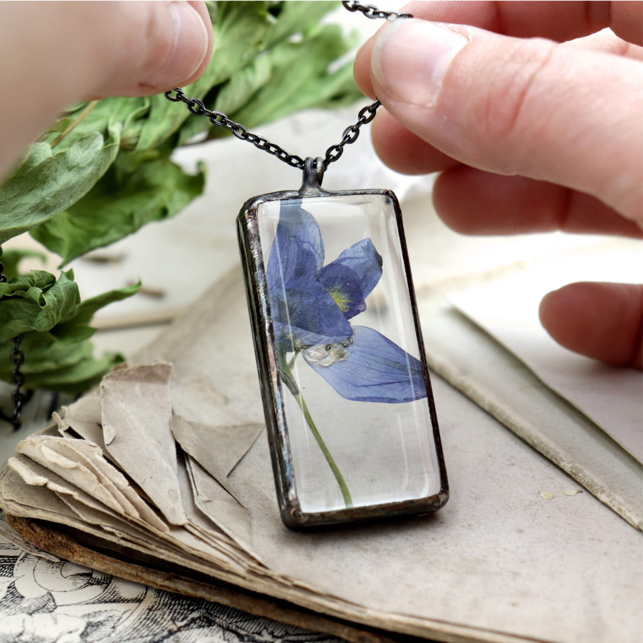 Hands holding a blue pressed flower in a rectangular glass soldered necklace