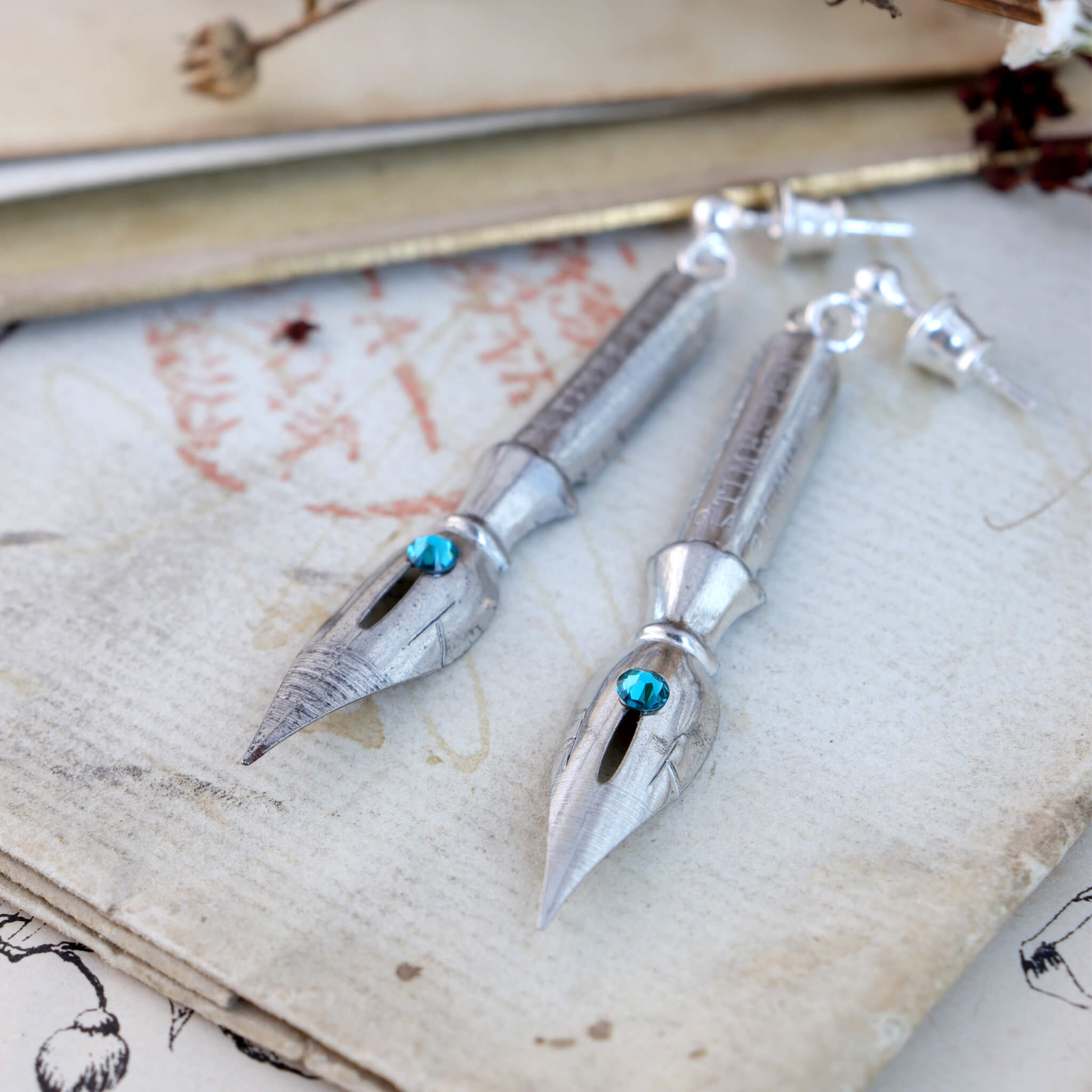 pen nib earrings with blue zircon birthstone crystals on them lying on vintage paper