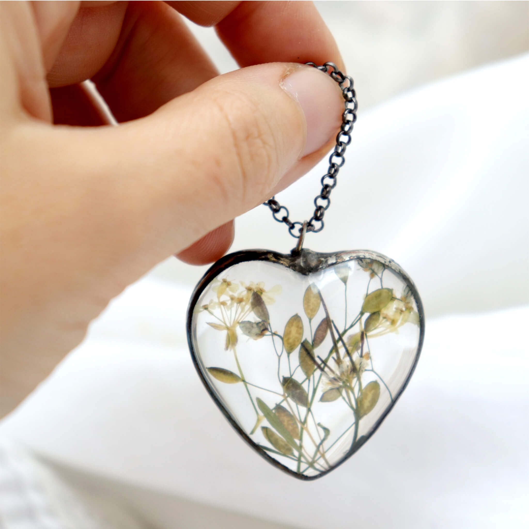 Hand holding heart shaped glass necklace with some plants inside it