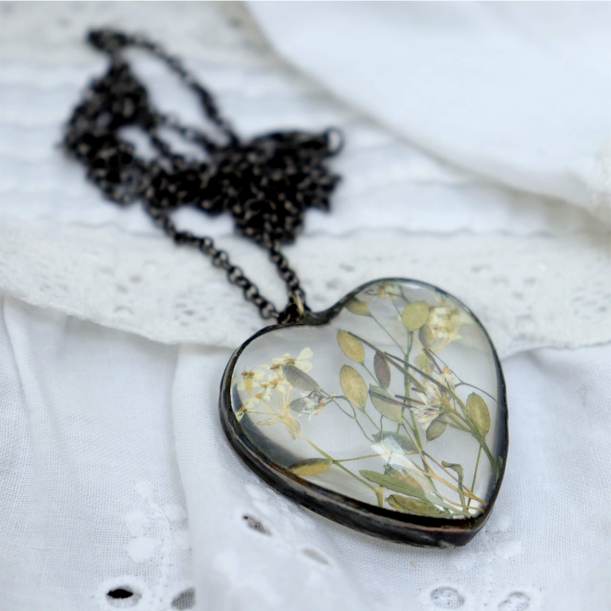 heart shaped glass necklace with some plants inside it lying on a white lacy fabric