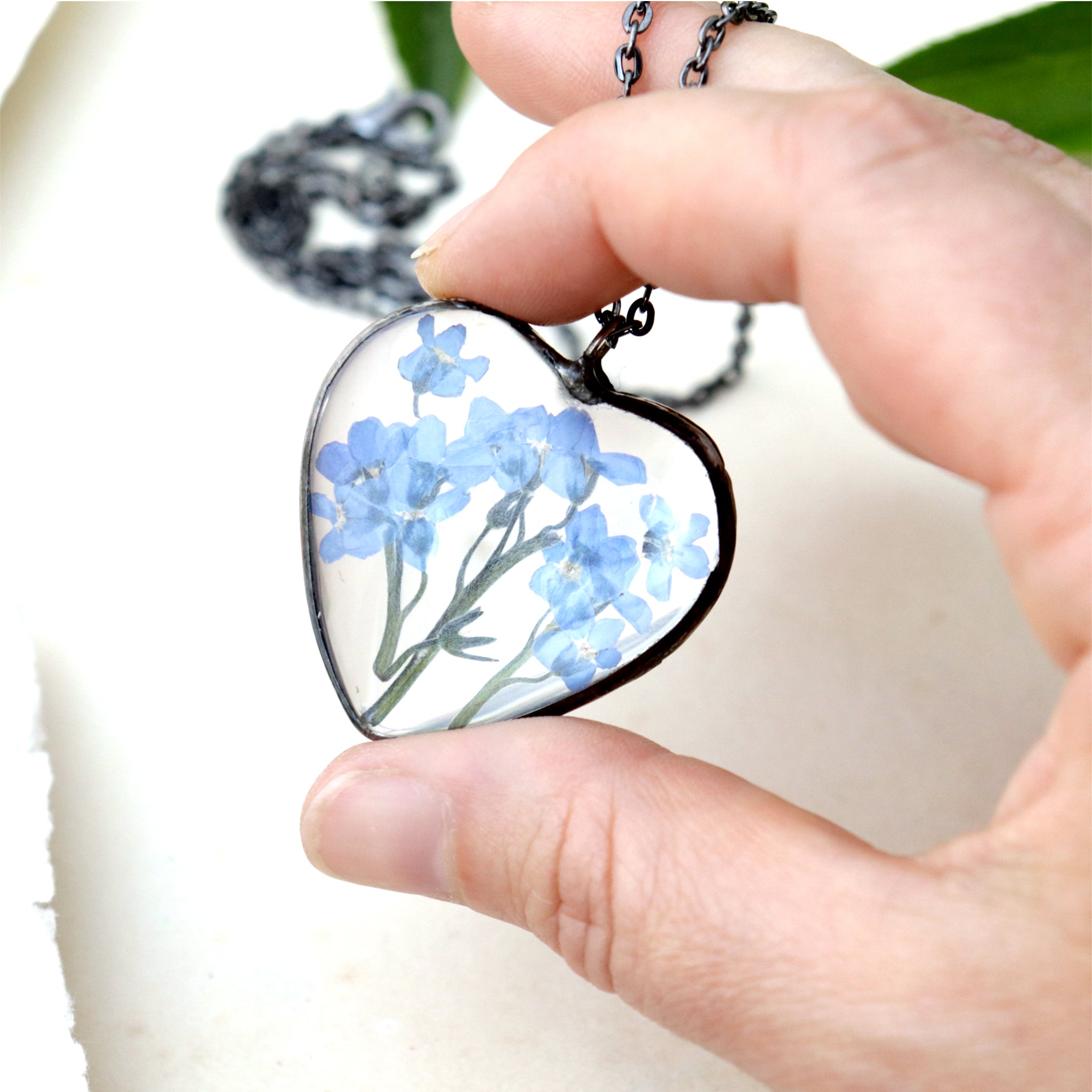 Forget-me-not heart shaped necklace being hold between thumb and index