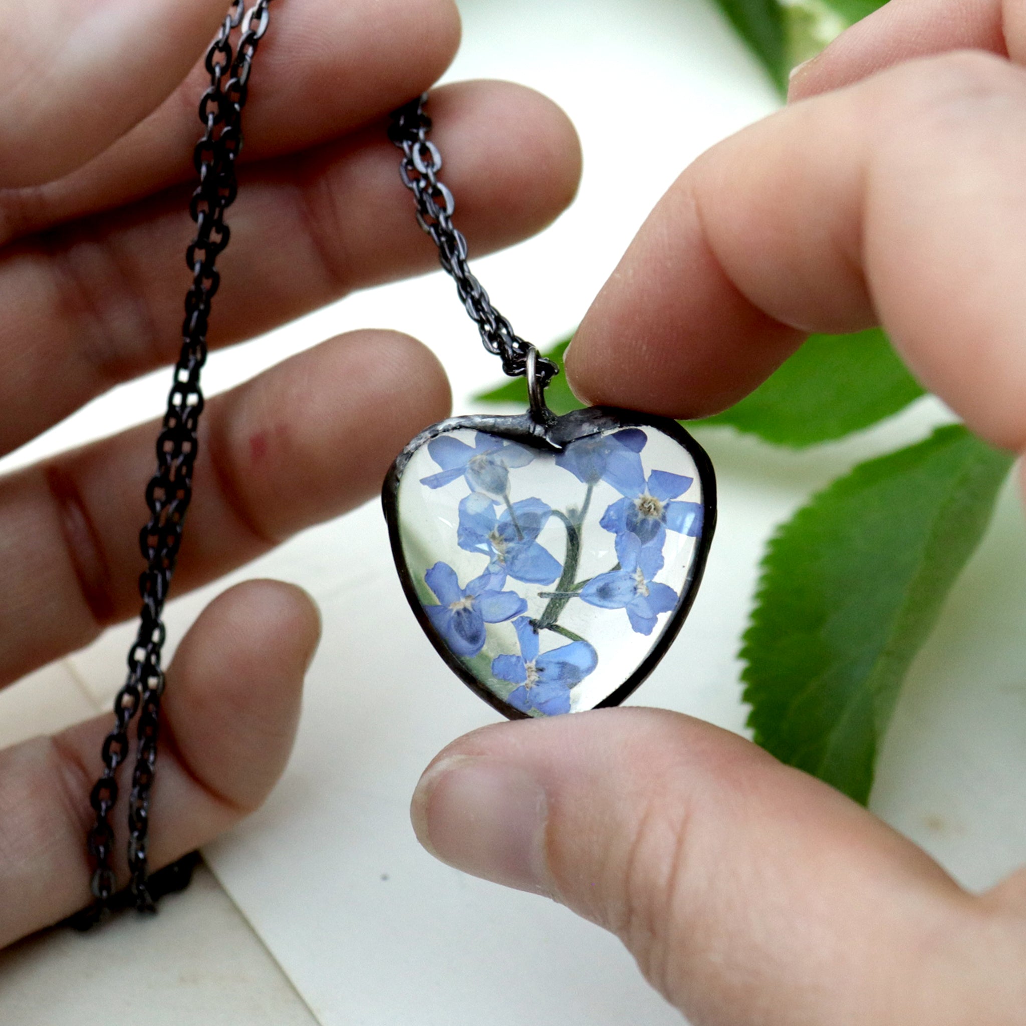 Forget-me-not heart shaped necklace being hold between thumb and index chain held by other hand