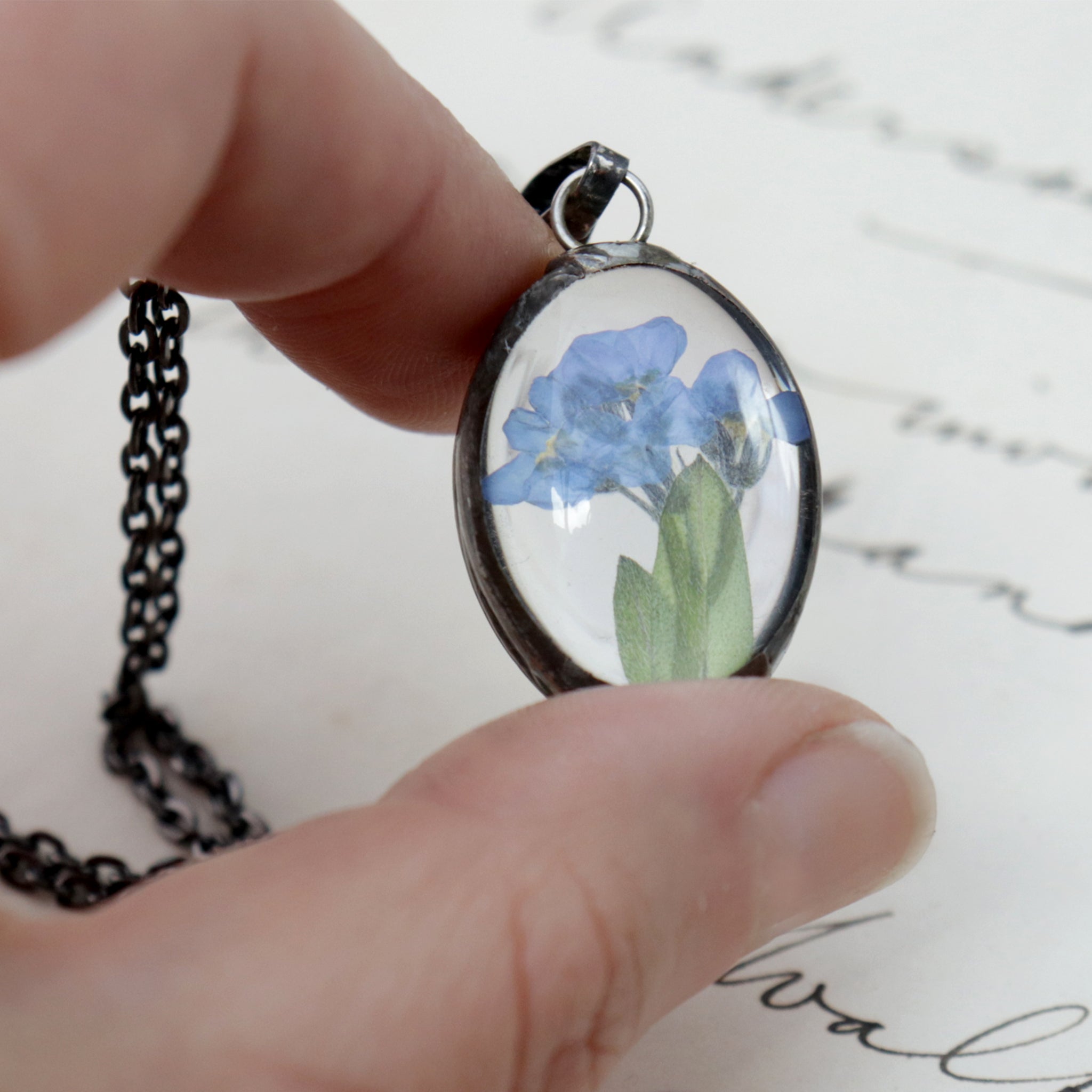 Forget-me-not oval little necklace being hold between thumb and index