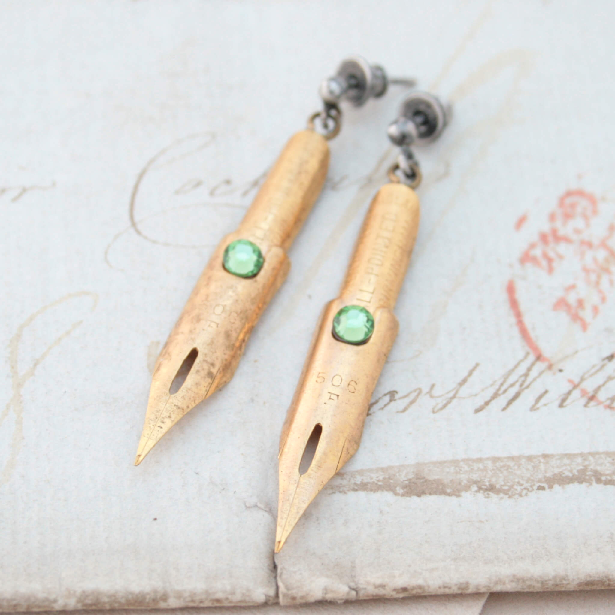 dark academia earrings made of gold coated pen nibs with peridot crystals lying on an old letter