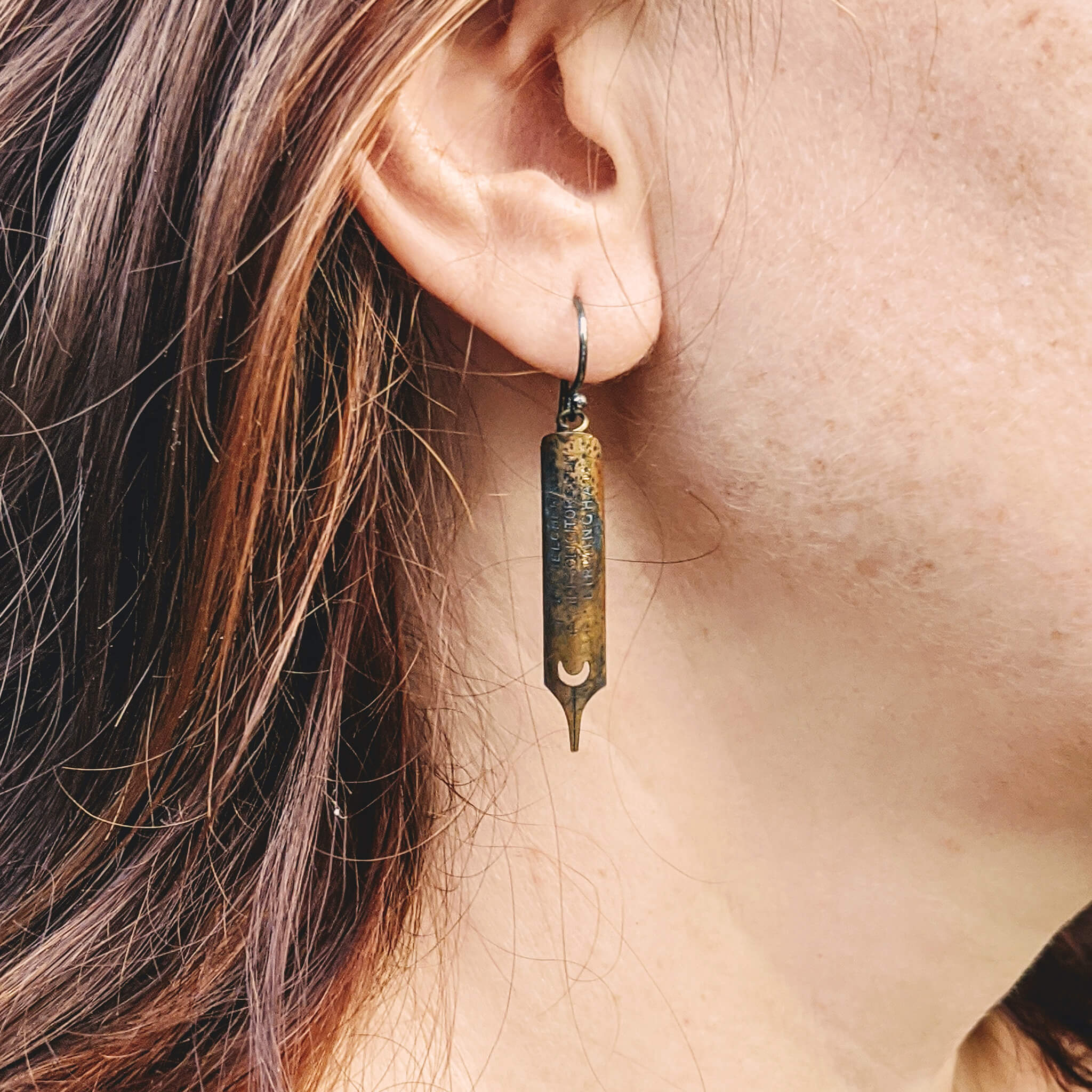 brunette woman's ear with antique gold pen nib earrings covered in patina