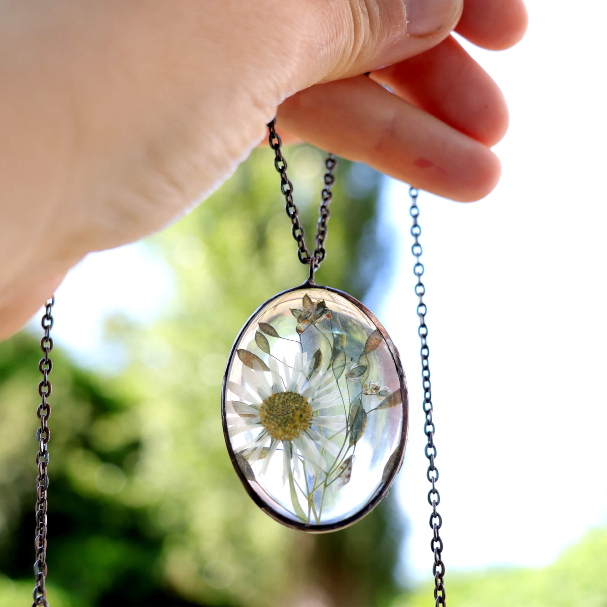 Hand holding oval glass soldered necklace with pressed daisy inside on a green background