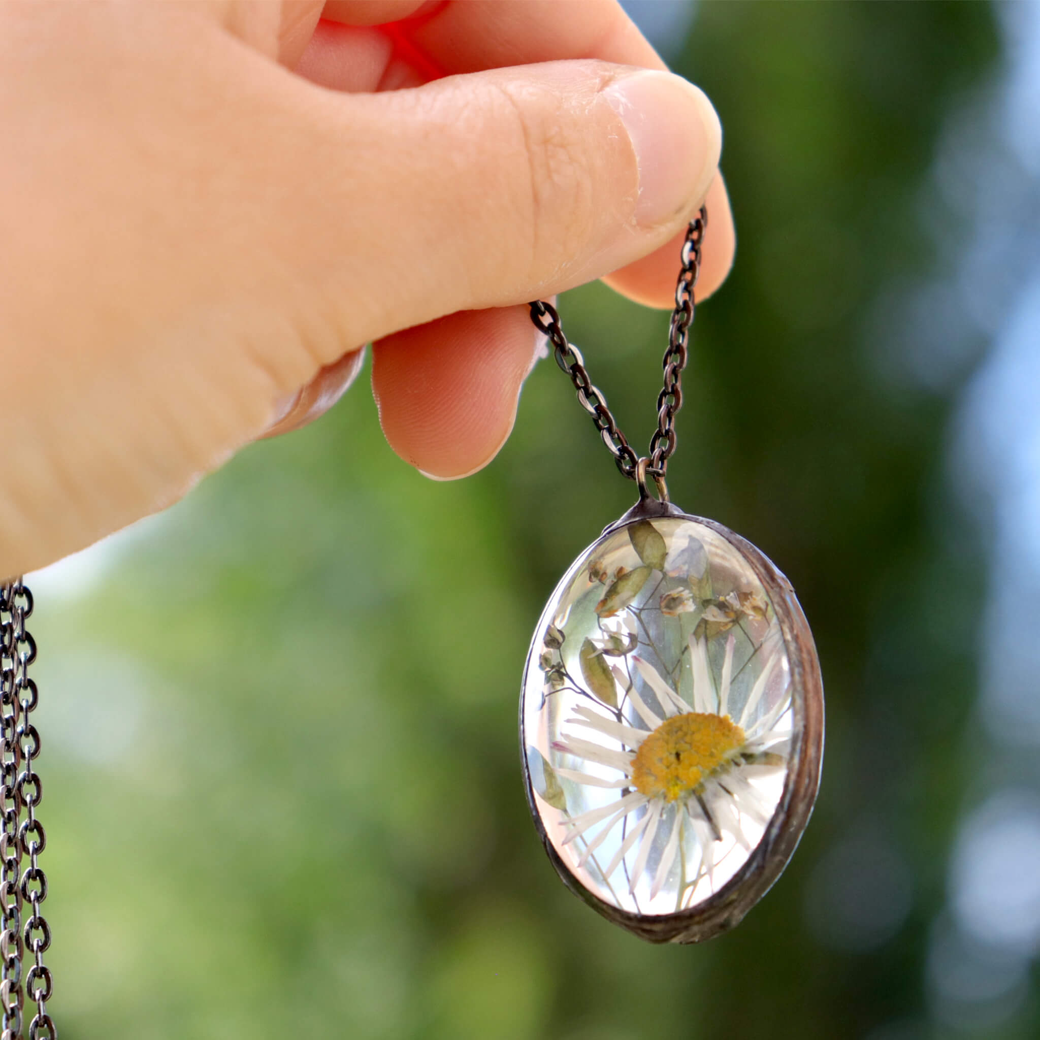 Hand holding oval glass soldered necklace with pressed daisy inside on a green background