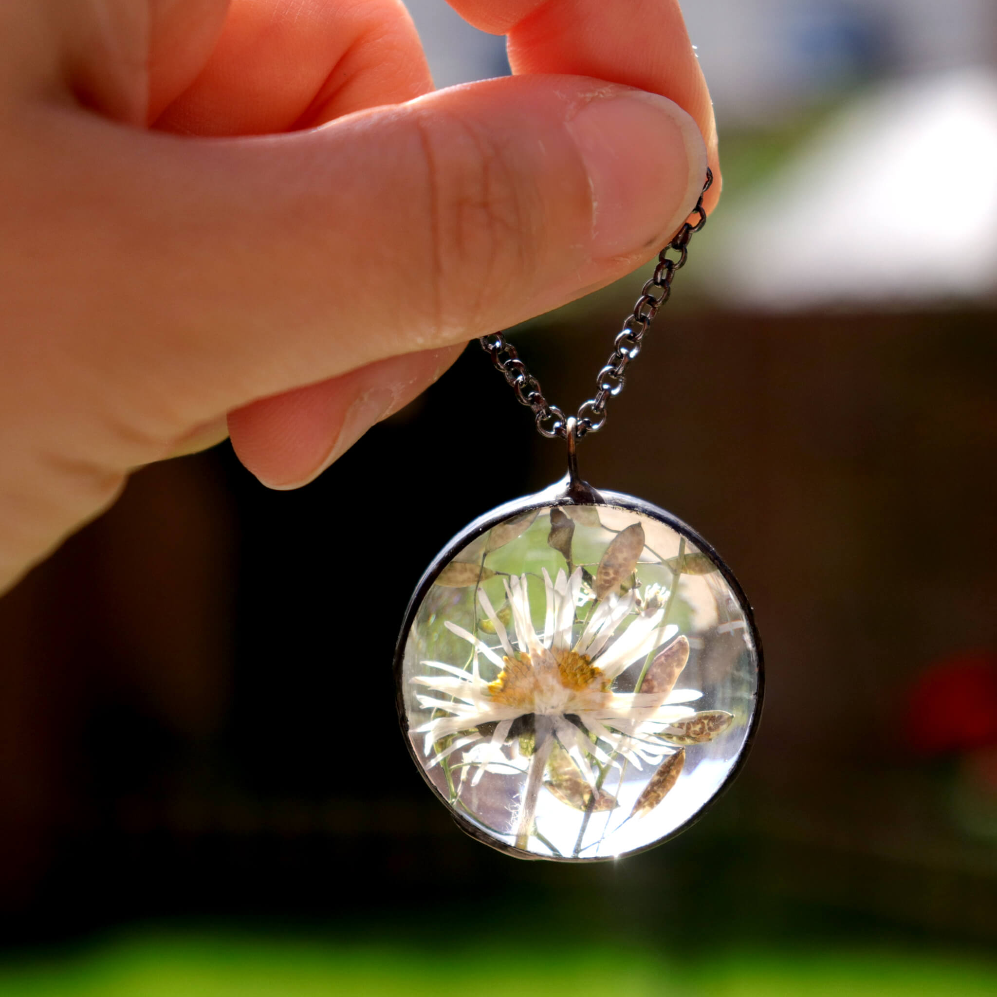 Hand holding round glass soldered necklace with pressed daisy inside on a dark background
