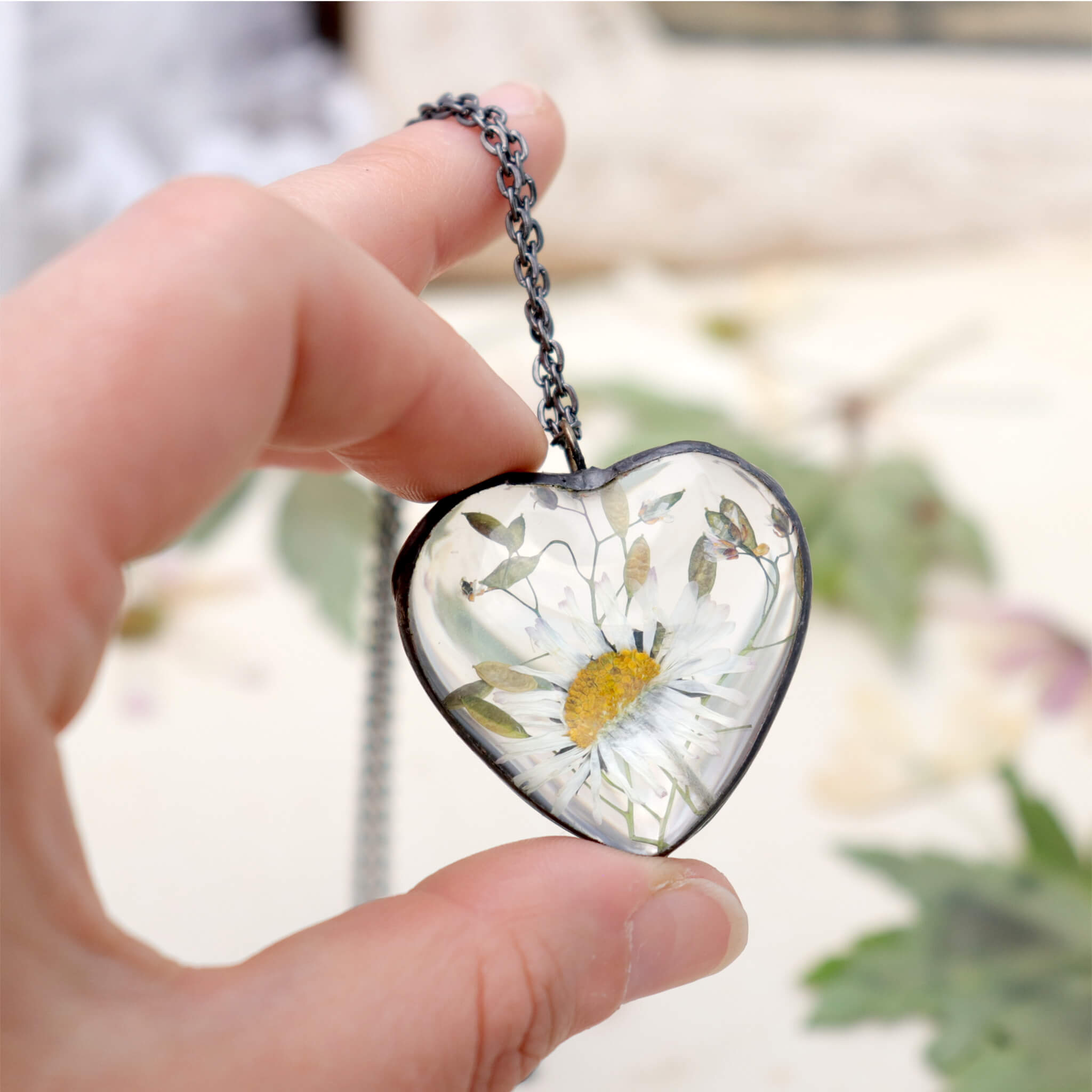 Hand holding heart shaped glass soldered necklace with pressed daisy inside on a pale background