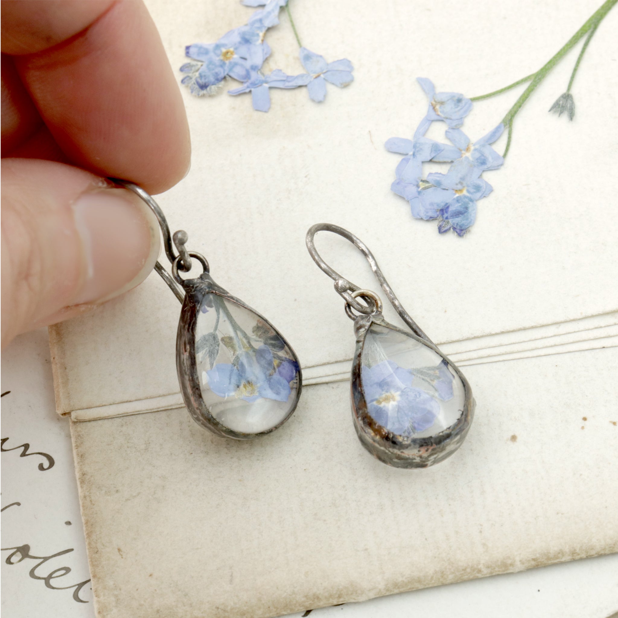 Teardrop shaped forget-me-not earrings lying on an old letter one being held in hand