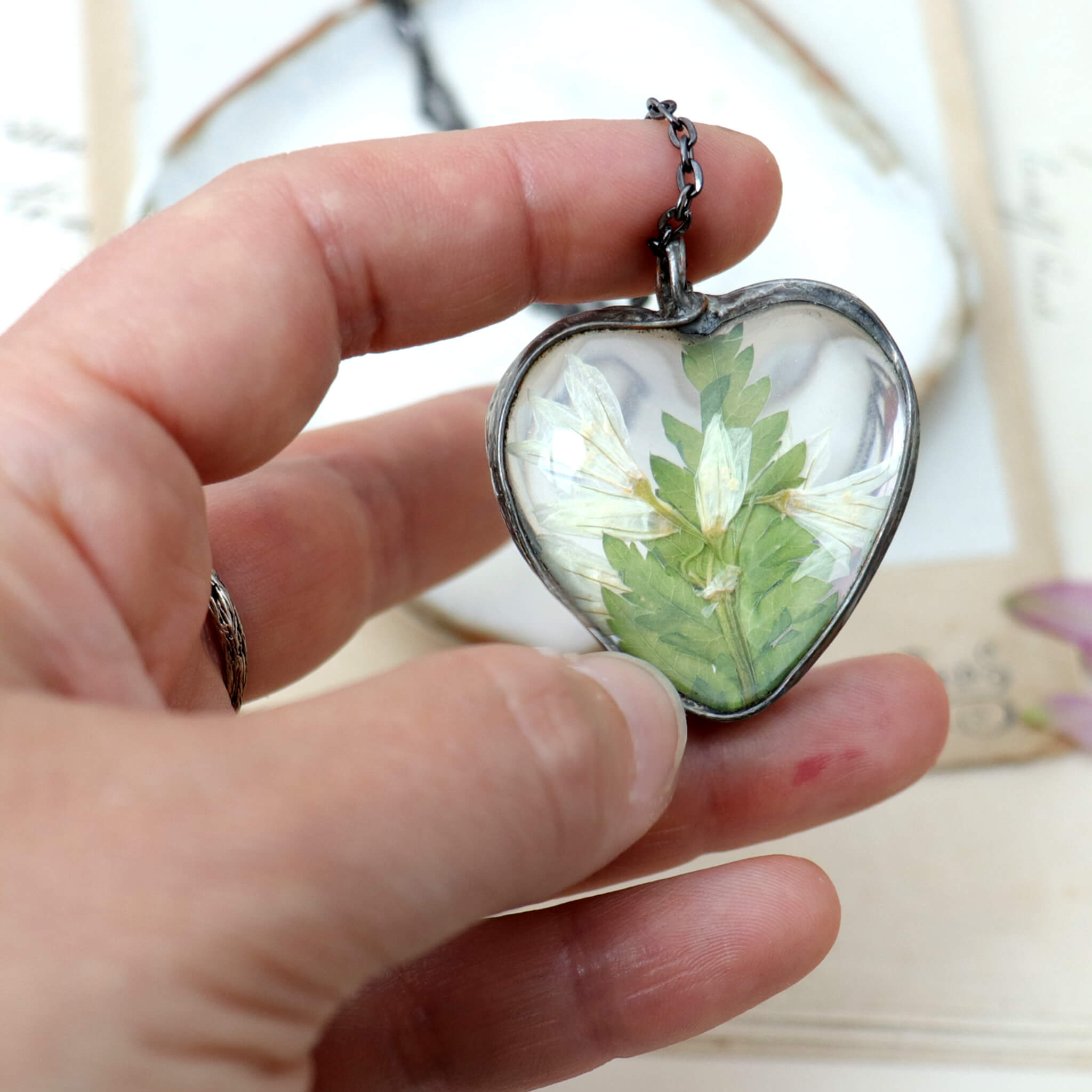 Hand holding heart shaped necklace with pressed flowers in focus blurry background