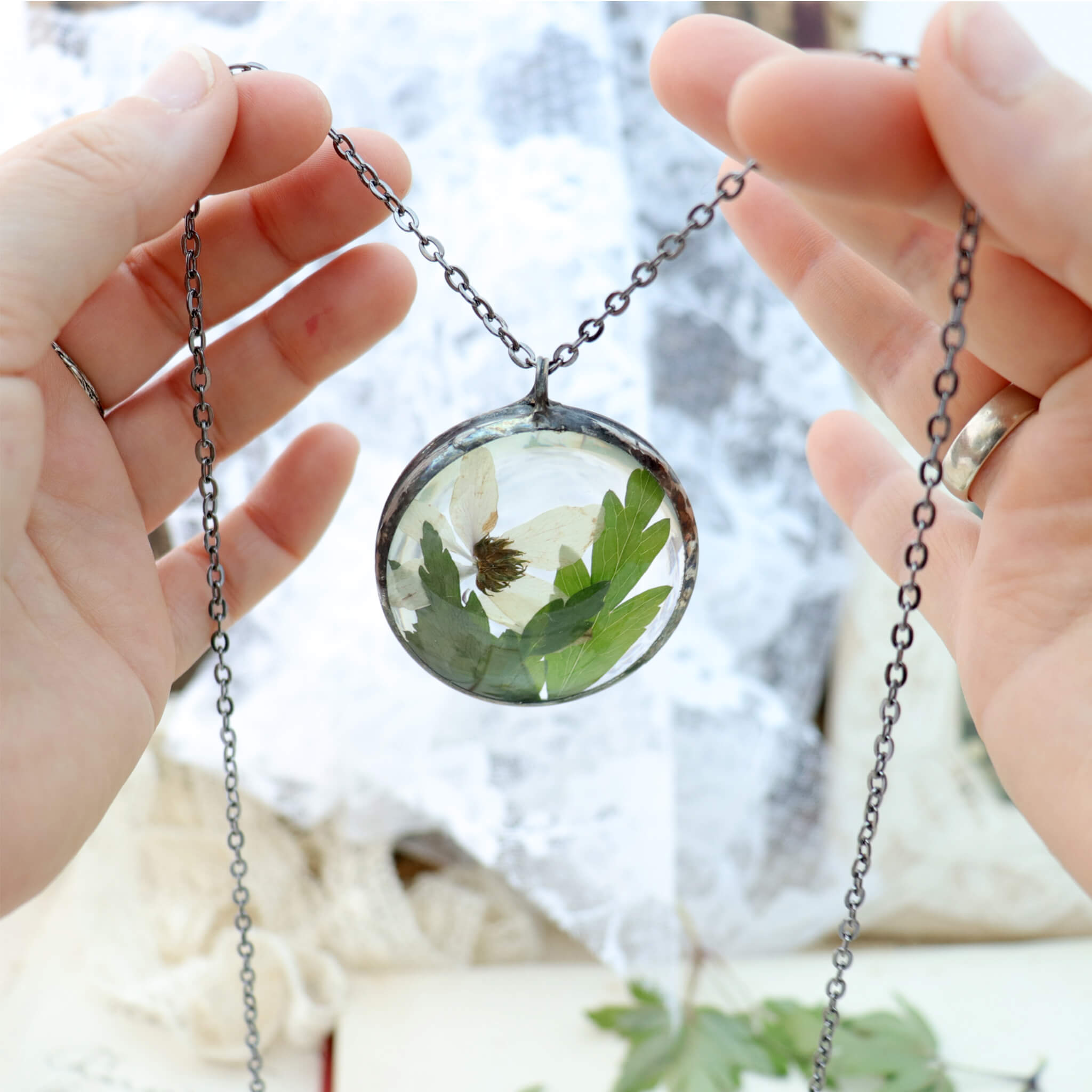 Hands holding round shaped necklace featuring pink pressed flowers with green leaves