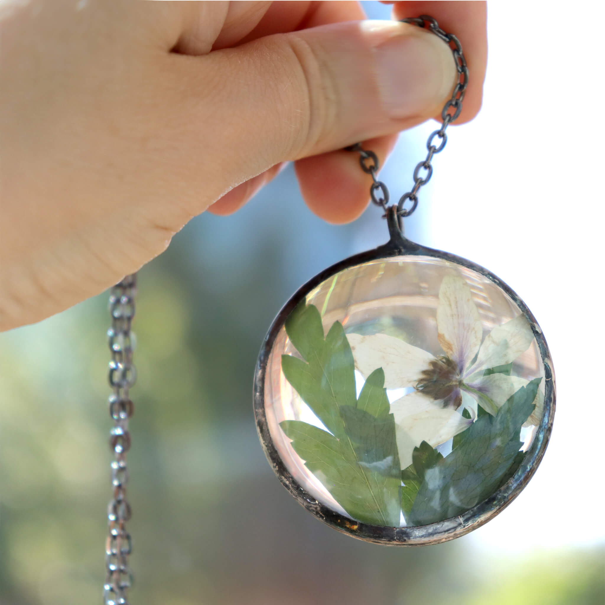 Hand holding round shaped necklace featuring pink pressed flowers with green leaves