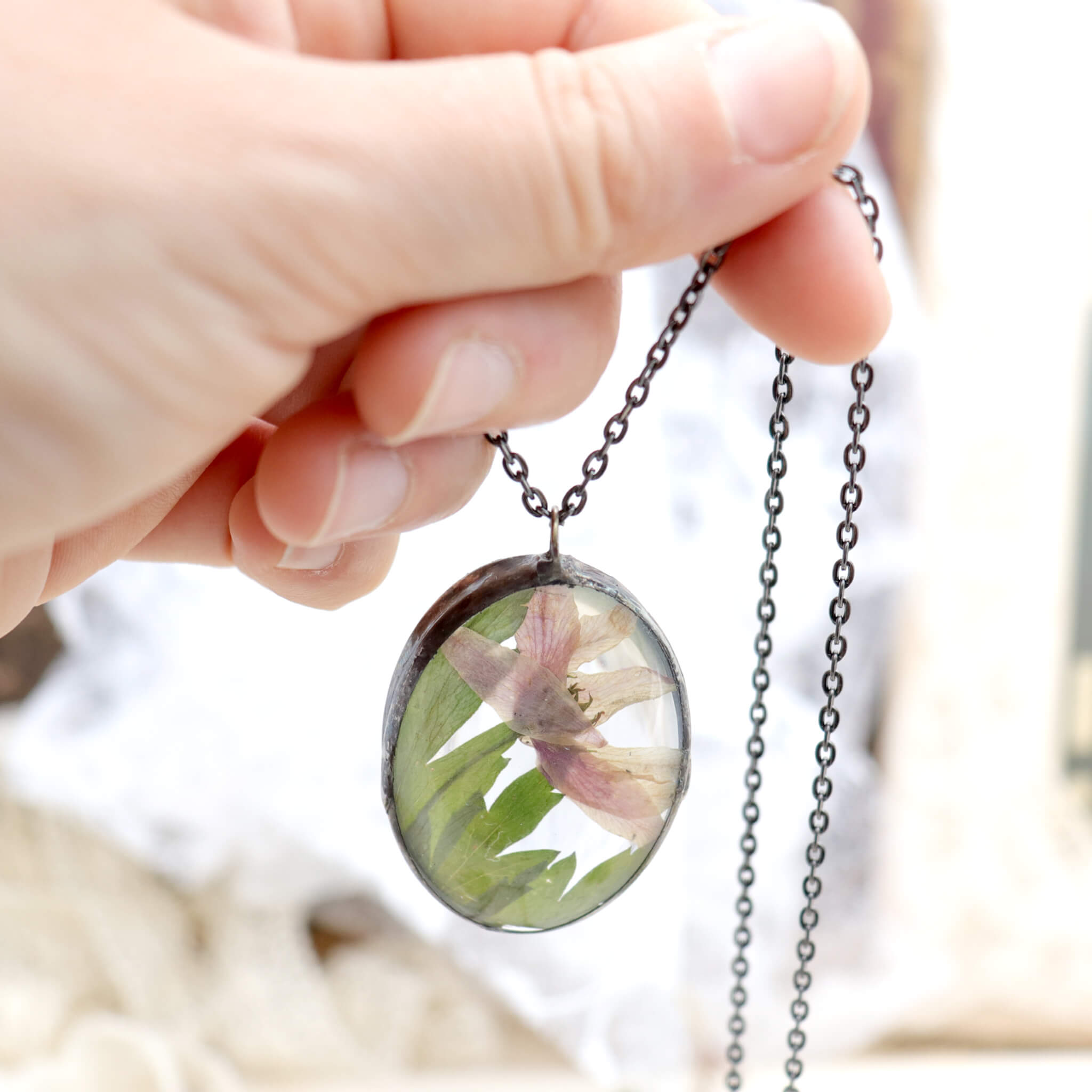 Hand holding oval shaped necklace featuring pink pressed flowers with green leaves