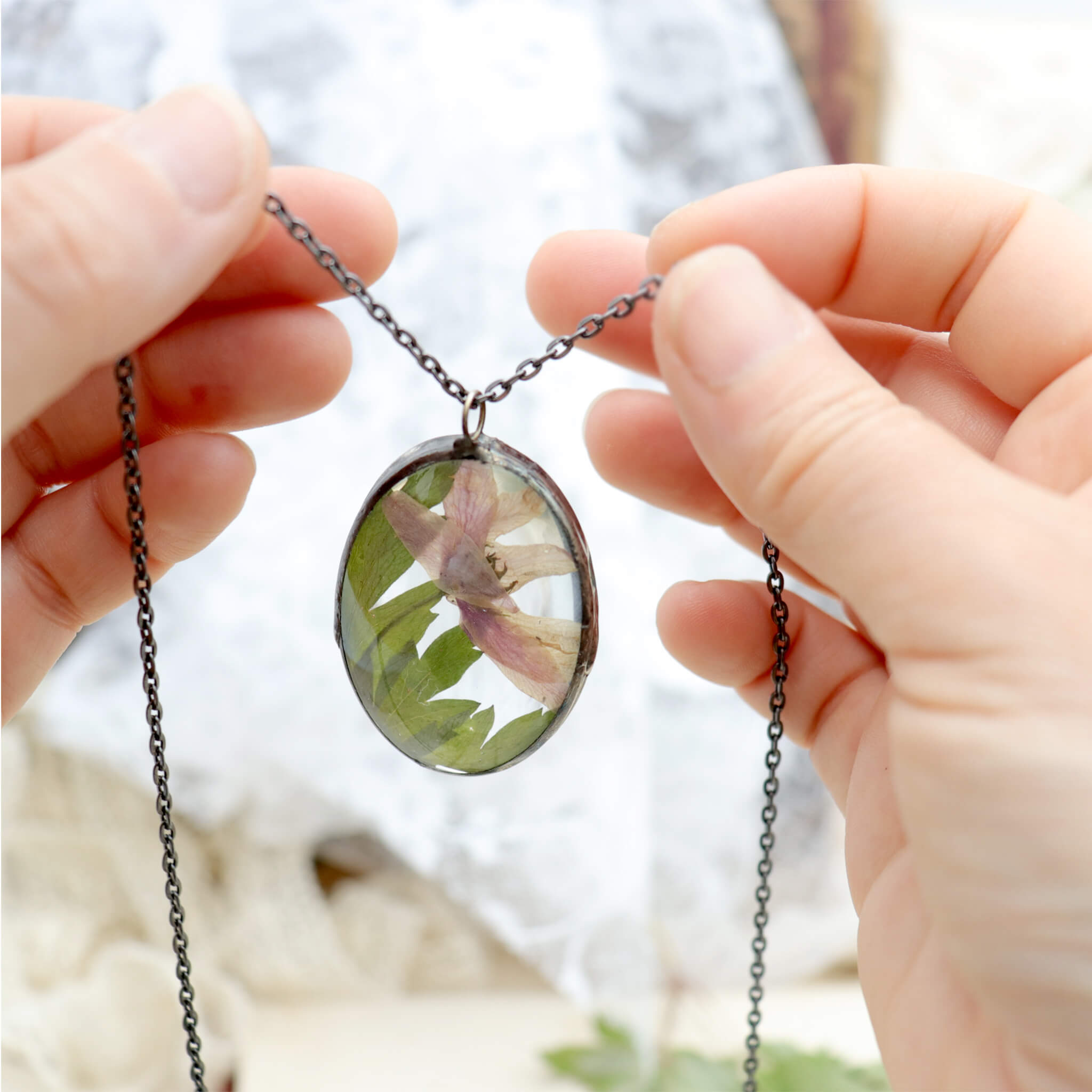 Hands holding oval shaped necklace featuring pink pressed flowers with green leaves